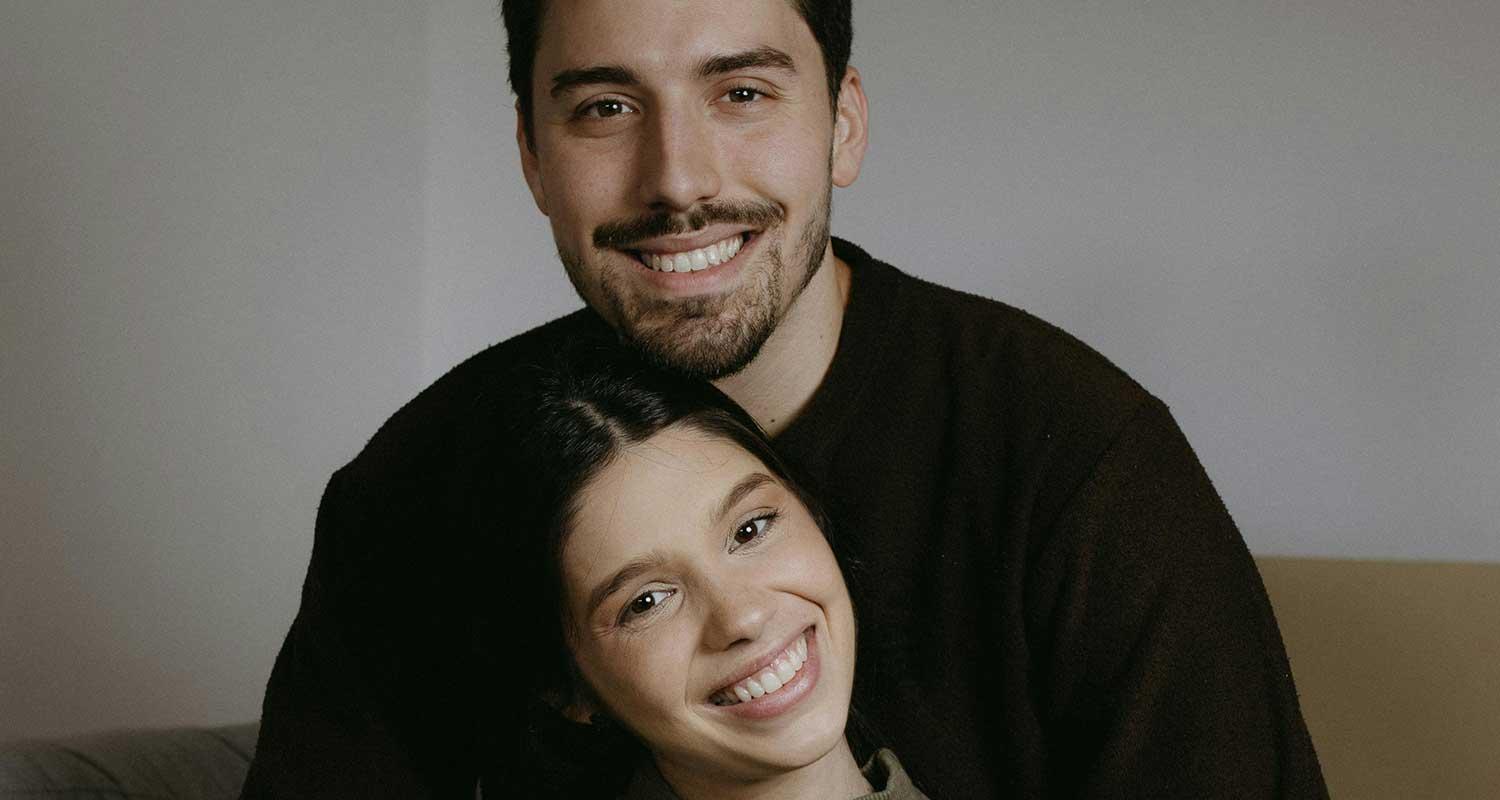 Ein sitzendes Paar. Beide lächeln. Veneers können helfen ein natürliches Lächeln wieder herzustellen, wenn etwa Zahnfehlstellungen, kleine Schäden und Verfärbungen das ästhetische Erscheinungsbild des Gebisses beeinträchtigt haben.